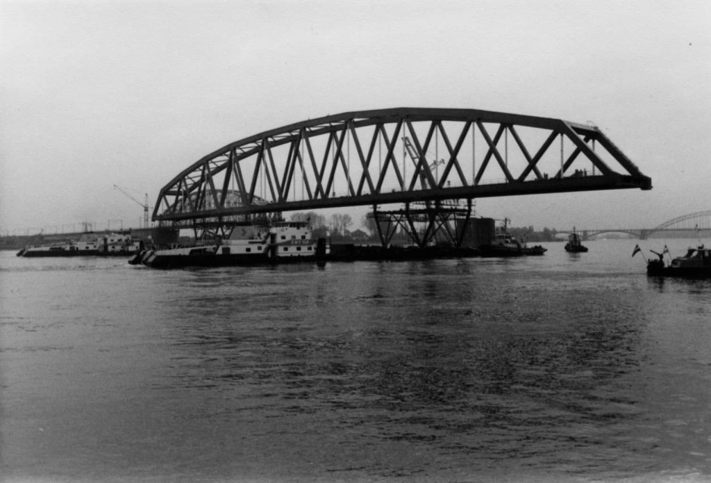 Bouw/modernisering van de spoorbrug. Vervanging van twee van de drie segmenten van de oude brug door een boog van 235 meter. Invaren van de nieuwe brug. De derde overspanning aan Lentse zijde zal, volgens draaiboek van de NS, in de tweede helft van 1984 vervangen worden door een uit drie delen bestaande betonnen aanloop, 8-5-1983 (J.J. van Ewijk via F88183 RAN CC0)