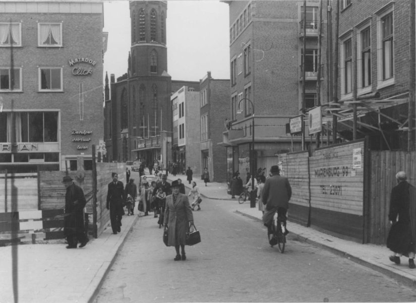 Gezien vanuit de Molenstraat. Rechts de bouw van het pand van Banketbakker W. van Loon (Molenstraat 3) en links daarvan het pand van Schoenhandel Van Haren (op de hoek met de Pauwelstraat) ; geheel links de bouw van het pand van Boekhandel Kloosterman, op de hoek met Plein 1944 ; in het midden de St. Dominicuskerk aan de Broerstraat met daarvoor de winkelpanden van o.a. het Warenhuis Firma A.A. Van der Borg en van Juwelier Firma Jac van Baal.