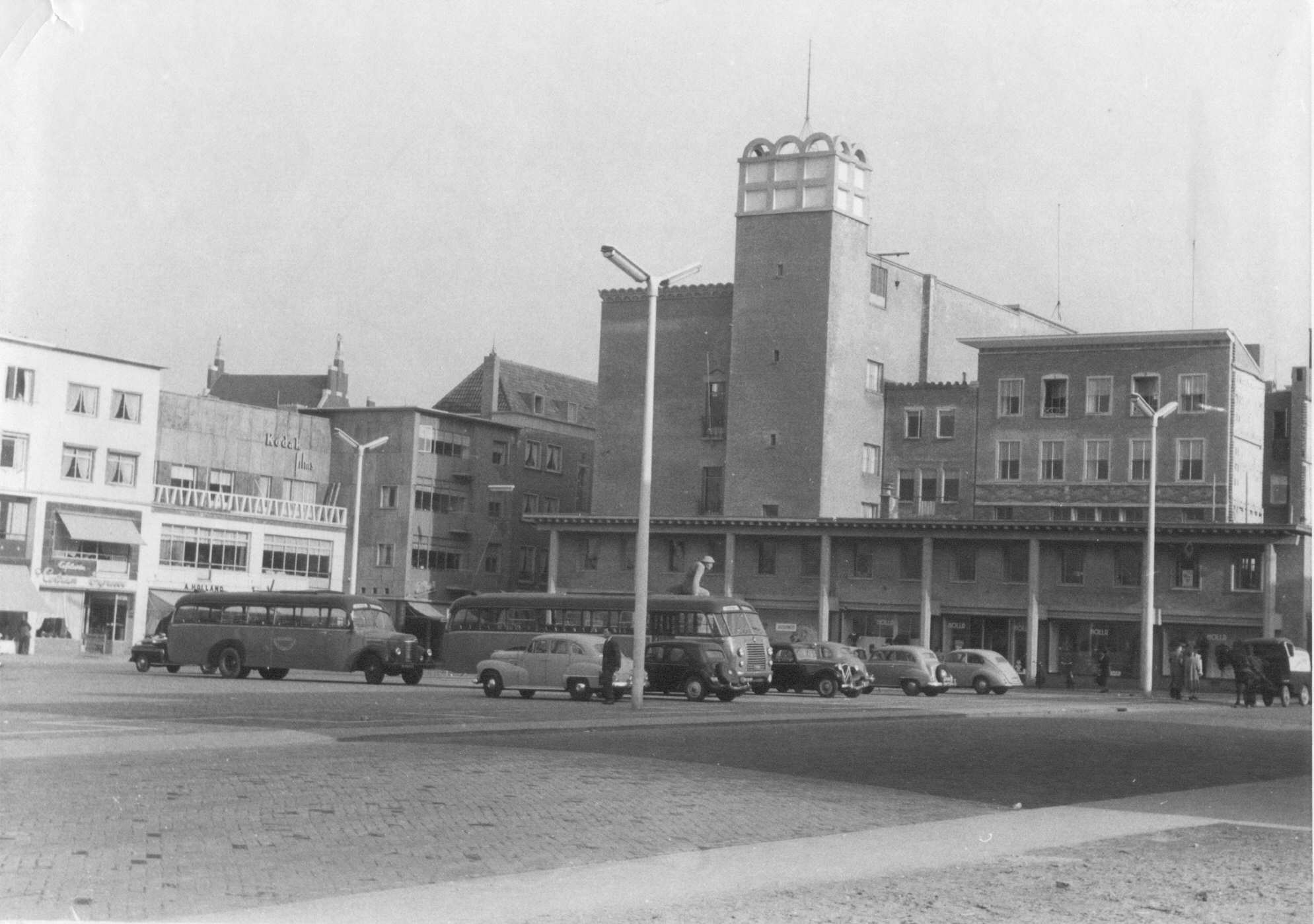 De oostzijde met het Kledingmagazijn van H.C. Holla (Plein 1944 nr. 152-153); links daarvan de noordzijde van Plein 1944 met v.l.n.r. Cafetaria Centrum Expresse, Schoenmagazijn A.J. Holland , Fotohandel A.M. Verweij en Parfumerie J.E. Albers. In het midden het pand van Van der Werff, 1953 (Commissariaat van Politie, Afd. Fotografie via F31812 RAN CC0) vd Werff architecten Pouderoyen Deur Molenstraat Plein 1944