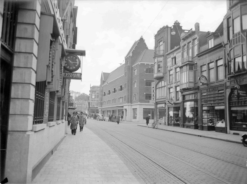 Manufacturenmagazijn Duives met op de de achtergrond het nieuwe complex; Straatbeeld zonder stadsschouwburg. Na veel gesteggel in de gemeenteraad werd op 14 april 1935 begonnen met de afbraak van de schouwburg. Een klein deel van het vrijkomende terrein werd benut om de Burchtstaat te verbreden. Het lukte pas geruime tijd later het terrein te verkopen aan de NV Handel en Exploitatie Mij. Zeekant uit Den Haag, die er naar een ontwerp van de architect L.D. Kuipers een complex met 7 winkels en 9 afzonderlijke bovenwoningen realiseerde. Links de Incasso Bank en rechts op de hoek met de Oude Stadsgracht de herenmode zaak van Duives. Het nieuwe complex was overigens geen lang leven beschoren, het werd verwoest tijdens gevechten in september 1944 door terugtrekkende en brandstichtende Duitsers. 1938-1939 (GN11015 RAN)