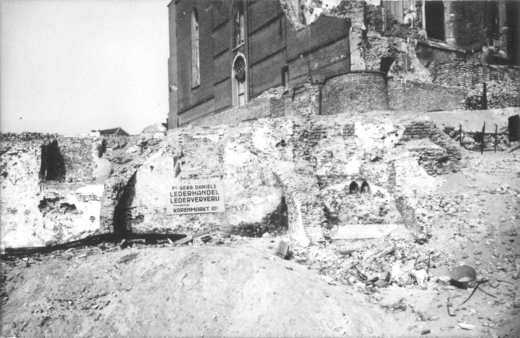 Het puin is geruimd op de hoek van de Stikke Hezelstraat met de Ganzenheuvel (links) en de Houtstraat (rechts) na het bombardement van 22 februari 1944; het bord verwijst de klanten van lederhandel Daniels naar de noodwinkel op de Korenmarkt. Nog net zichtbaar de torenvoet van de Sint Stevenskerk, 2/1944-12/1944 (F45999 RAN)