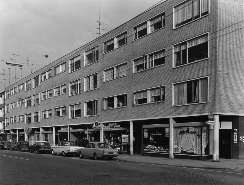 Het nieuwgebouwde woon-winkelcomplex aan de oostzijde van de Bloemerstraat (nrs.: 2 t/m 48). met o.a. de Milord-bar (nr.: 2), een bakkerswinkel van de Roding (nr.: 4) , Autorijschool Brugmans (nr.: 12-14). Kledingmagazijn Amelink (nr.: 16-18) en Chinees Restaurant Hong Kong (nr.: 30), 1961-1964 (J.H. ten Have via F39115 RAN CCBYSA Auteursrechthouder Hans en Tijs ten Have)