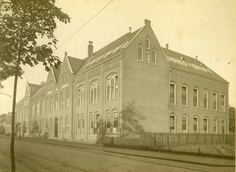De eerste Nijmeegse Ambachtsschool, opgericht door de Vereeniging Ambachtsonderwijs voor Nijmegen in 1901 en (aansluitend) gebouwd in opdracht van het gemeentebestuur naar ontwerp van gemeentearchitect ir. Jan Jacob Weve. Op de voorgrond de rails van de kolentram voor het vervoer van de steenkool van het station naar de Centrale aan de Waalkade, Nieuwe Marktstraat 12, 1919 (Born via F38489 RAN)
