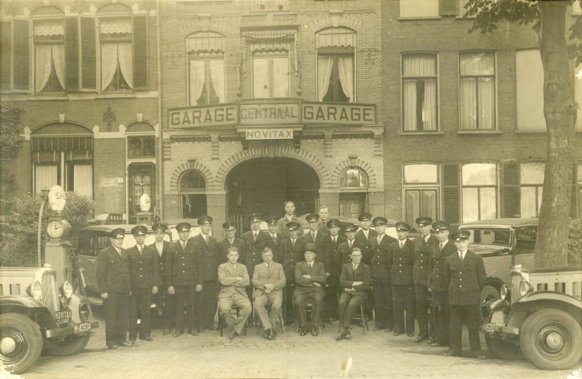 Eigenaar J.H.W. van Bon (met hoed) van Autobedrijf Van Bon poseert met zijn drie zoons Antoon (rechts), Herman en Theo (links), de chauffeurs van taxi-onderneming Novitax en personeelsleden van de Centraal-Garage voor de ingang. Op de achtergrond rechts (auto)monteur Gijs Nas, Nieuwe Markt 8. 1934 (F26525 RAN Auteursrechthouder B. van Bon)
