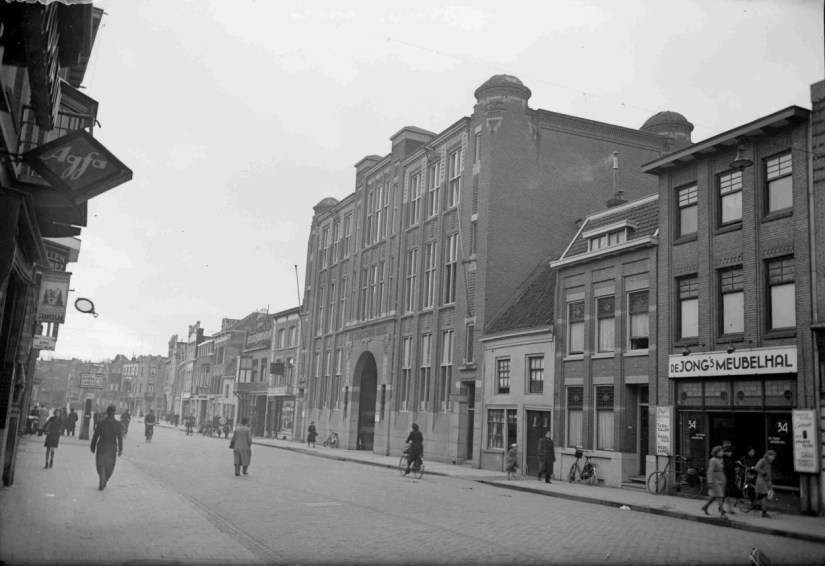 Tussen de lage bebouwing troont het gebouw van de Twentsche Bank, in 1921 gereedgekomen naar een ontwerp van Oscar Leeuw. Links van de bank het Eijsinkhuis (motor- en rijwielhandel) en daarnaast Heineken's Bierbottelarijen; aan de rechterzijde de stadsboederij van R.J. Faber (melkhandel), de mode vakschool en de Jong's meubelhal. Boven de Bank kantoren onder andere van de N.V. Verenigde Draadfabriek, Hertogstraat, 2/1/1942 (GN11104 RAN)